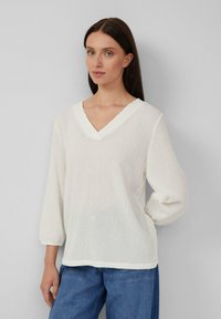 Vrouw draagt een witte, geweven V-hals blouse met 3/4 mouwen en blauwe jeans, staande tegen een lichtgrijze achtergrond.
