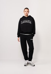 Czarny bawełniany hoodie z logo "LAMANIA", z kieszenią z przodu, połączony z dopasowanymi czarnymi spodniami dresowymi. Model nosi srebrne sneakersy.
