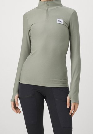 Vrouw draagt een lichtgroene geribbelde longsleeve met halve rits en logopatch, gecombineerd met een zwarte broek met voorzakken.