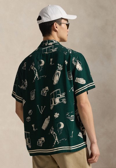 Mężczyzna w ciemnozielonej koszulce z krótkim rękawem, ozdobionej białymi ilustracjami o tematyce golfowej oraz białymi i beżowymi paskami na krawędziach, beżowych spodniach, białej czapce i okularach przeciwsłonecznych.