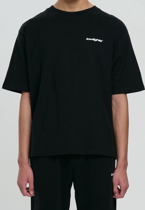 Person iført en løs sort t-shirt med "lowlights" logo i hvid på venstre bryst, kombineret med sorte bukser.
