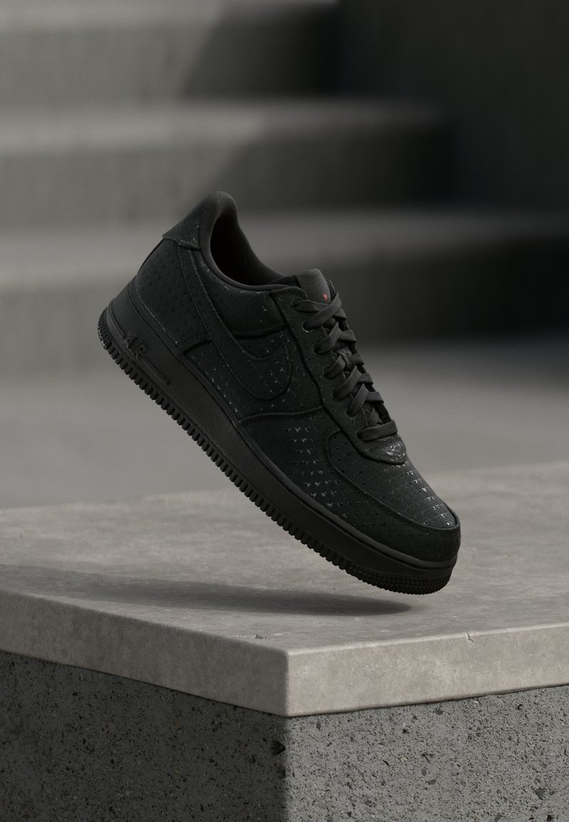 Μαύρο αθλητικό παπούτσι Nike με διάτρητες λεπτομέρειες αιωρούμενο πάνω από μια τσιμεντένια άκρη με φόντο ένα θολό γκρι φόντο.