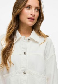 Chaqueta de mezclilla blanca con cuello amplio, botones frontales y dos bolsillos en el pecho. Textura suave con detalles de costura visibles.