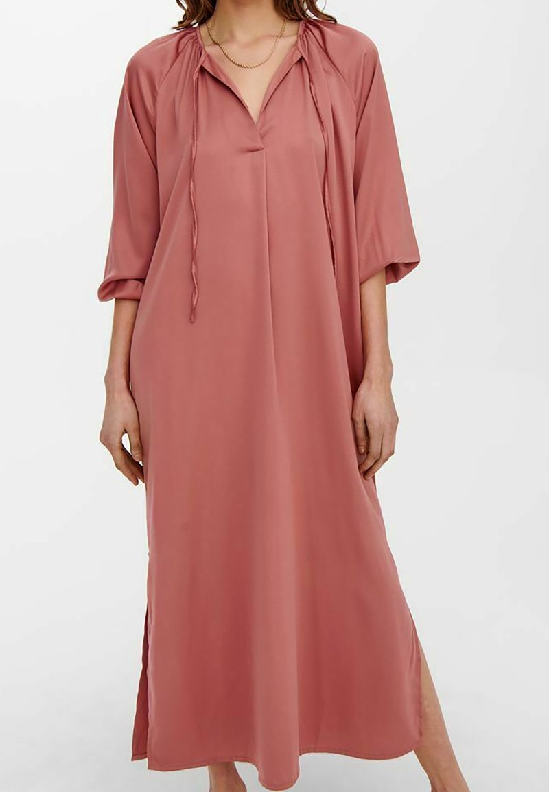 Femme portant une longue robe ample rose poudré avec des manches trois-quarts, des fentes latérales, un col en V et des liens décoratifs au niveau de l'encolure.