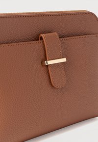 Καφέ πορτοφόλι από δερματίνη με υφή, με μεταλλικό κούμπωμα σε χρυσή απόχρωση και φερμουάρ στην κορυφή, εμφανιζόμενο σε απλό λευκό φόντο.