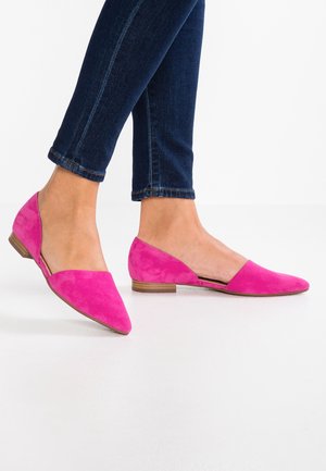 Persoon draagt donkerblauwe spijkerbroek en felroze platte schoenen met puntige tenen op een witte vloer.