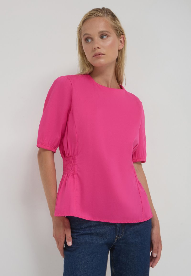 Donna con una blusa rosa brillante con maniche a sbuffo e dettagli arricciati in vita, abbinata a jeans blu scuro, su uno sfondo neutro.