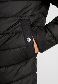 Svart puffjacka med sydda horisontella paneler, en ficka med dragkedja och silverdetaljer, samt en mjuk texturerad yta.