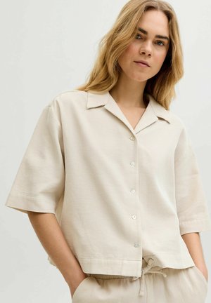 Donna bionda che indossa una camicia beige a maniche corte, ampia e abbottonata, e pantaloni abbinati con coulisse, con le mani nelle tasche, sfondo neutro.