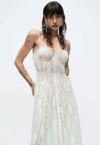 Žena s tmavými vlasmi a tetovaniami, ktorá nosí dlhé priehľadné biele kvetinové čipkované šaty a visiace náušnice na jednoduchom svetlom pozadí.