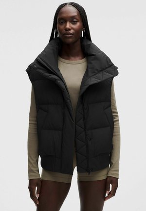 Donna con lunghe trecce che indossa un gilet imbottito nero senza maniche sopra una maglietta beige a maniche lunghe e pantaloncini abbinati su uno sfondo semplice.