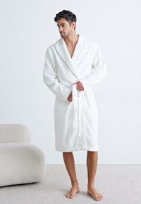 Biely bavlnený župan so šálovým golierom, viazaný v páse; textúrovaná látka a dĺžka po polovicu lýtok. Model bez obuvi stojí na svetlom koberci.