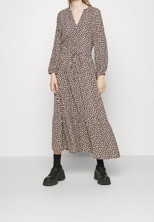 Frau trägt ein langärmliges, knöchellanges Kleid mit abstraktem beige-dunklem Muster, kombiniert mit schwarzen Socken und klobigen schwarzen Schuhen.