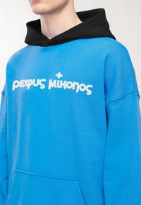Homme portant un sweat à capuche bleu avec un texte blanc et une capuche noire, montré du cou jusqu'au milieu du torse sur un fond uni.