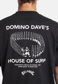Black cotton t-shirt featuring white graphic with surfboards, cartoon character, and text "DOMINO DAVE'S HOUSE OF SURF" in bold font.