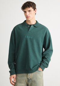 Tumes roheline pusak, millel on polo kaelus, kaelas nööp ja rinnal tikand "HUGO" logo. Pehme tekstuur.