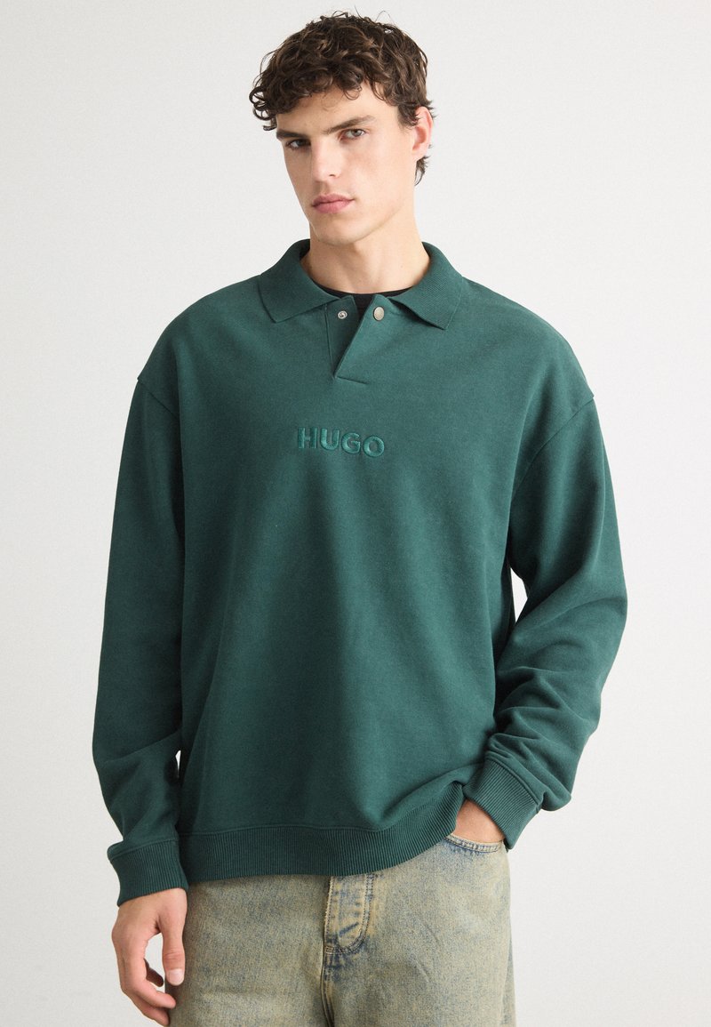 Tumes roheline pusak, millel on polo kaelus, kaelas nööp ja rinnal tikand "HUGO" logo. Pehme tekstuur.