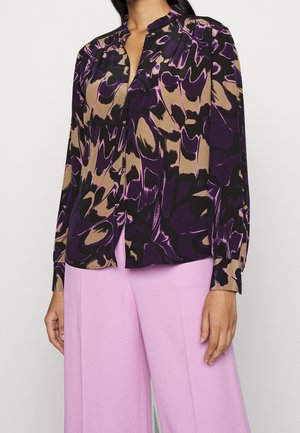 Kvinde iført en langærmet bluse med abstrakt mønster i lilla, sort og tan samt lyse lilla bukser med vide ben.