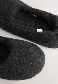 Pantofole in feltro nero con una texture morbida, forma arrotondata e fiocchi d'argento. Etichetta visibile con la scritta "NEXT".