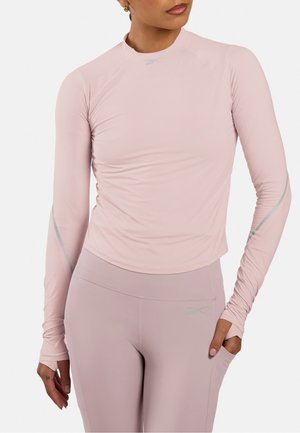 Frau trägt hell mauve eng anliegendes langärmeliges Sportoberteil und passende Leggings mit hoher Taille, eine Hand in der Tasche, einfarbiger Hintergrund.