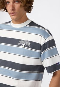 Jonge man die een blauwe, witte en grijze gestreepte Champion t-shirt draagt met een logo op de borst en de mouw, met een lichte draai naar links.