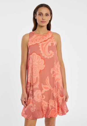 Frau trägt ärmelloses korallenfarbenes Kleid mit großen hellrosa Blumenmustern, steht vor weißem Hintergrund.