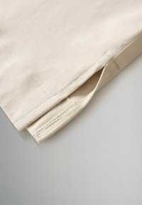 Gros plan sur un ourlet en tissu beige avec une fente latérale visible et une couture détaillée sur fond gris clair.