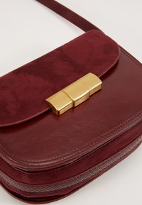 Borsa a tracolla in pelle bordeaux con patta in camoscio, chiusura in metallo dorato e dettagli testurizzati lungo i lati. Finiture lisce e opache.