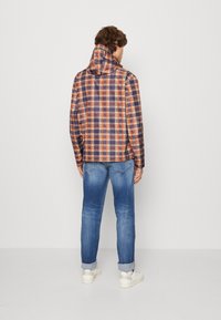Mand iført en orange og marineblå ternet hættetrøje, op-rullede blå jeans, hvide sokker og hvide sneakers, står vendt væk mod en ensfarvet baggrund.