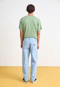 T-shirt in cotone verde chiaro con maniche corte, abbinato a jeans in denim azzurro a gamba dritta con tasche posteriori e un'etichetta marrone.