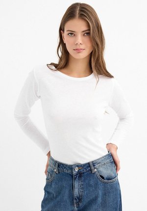 Camisa blanca de manga larga hecha de una tela suave, con un escote redondeado. Combinada con unos jeans azules de cintura alta. Diseño simple y limpio.