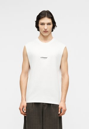 Ung mann med kort, mørkt hår iført hvit ermeløs T-skjorte med teksten "JJ Studios" og vide, mørke bukser, stående foran en ensfarget bakgrunn.