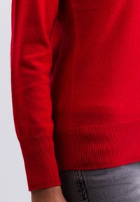 Röd stickad tröja med en slät textur, ribbade ärmslut och nederkant. Har en figurnära design som formar sig efter kroppen.