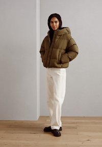 Doudoune vert olive avec capuche, dotée d'une fermeture à zip et d'une texture matelassée. Associée à un pantalon blanc à jambes larges et des chaussures noires.