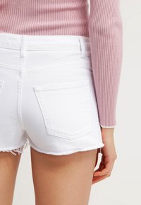 Pantalones cortos de mezclilla blancos con bordes desgastados y dos bolsillos traseros, combinados con un top rosa de manga larga acanalado.