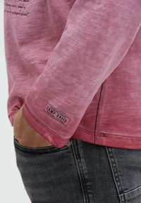 Long-sleeve shirt in light pink fabric with a textured finish. Features a logo tag on the bottom and visible stitching along the seams.