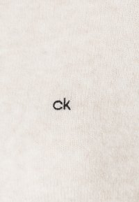 Gros plan sur un tissu beige avec un petit logo brodé "ck" noir centré sur la surface texturée.