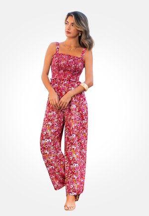 Femme portant une combinaison rose à motifs floraux, sans manches, à jambes larges, avec des talons, regardant sur le côté, sur un fond blanc uni.