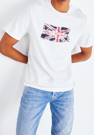 Mężczyzna w białej koszulce z przetartą flagą Wielkiej Brytanii i logo "Pepe Jeans", w połączeniu z jasnoniebieskimi przetartymi dżinsami.