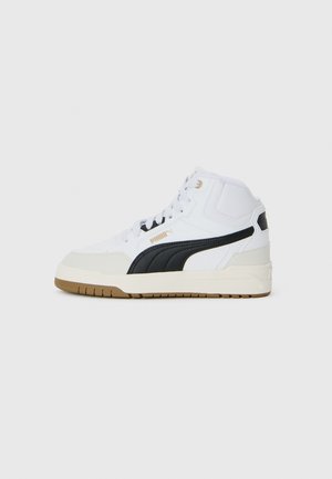 Baskets blanches mi-hautes avec une bande noire Puma, un embout beige, une semelle en gomme et des lacets blancs, sur un fond gris uni.