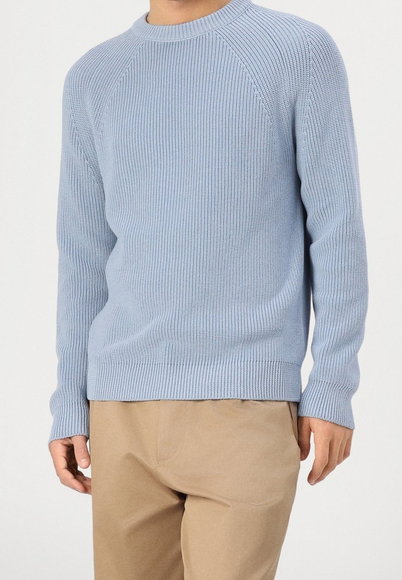 Light blue ribbed sweater with a round neck, long raglan sleeves, and a textured finish. Paired with beige pants, all against a white background.
