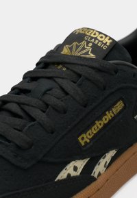 Baskets noires Reebok Classic avec logo doré, lacets texturés et bande latérale blanche tachetée sur semelle marron.