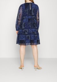 Marineblaues Kleid mit Patchwork-Muster, transparenten langen Ärmeln, mehrlagigem Rock und Schlüsselloch-Detail am Rücken, kombiniert mit hellen, römersandalenartigen Absätzen.