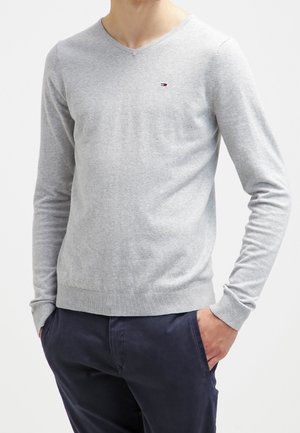 Homme portant un pull à col en V gris clair avec un petit logo sur la poitrine et un pantalon bleu marine foncé, les mains dans les poches.