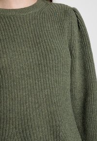 Zielony sweter dziany o prążkowanej fakturze, okrągłym dekolcie i delikatnie bufiastych rękawach. Materiał wydaje się miękki z lekko zróżnicowanym kolorem.