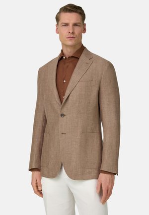 Man draagt een beige visgraatblazer over een bruin overhemd met knopen en witte broek, staand tegen een eenvoudige lichte achtergrond.
