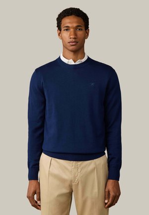 Joven con un jersey azul marino sobre una camisa blanca con cuello y pantalones beige, de pie frente a un fondo liso y claro.