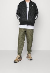 Svart quiltad bomberjacka med vita ränder, olivgröna quiltade byxor och bruna sneakers med camouflage-mönster, buren över en vit skjorta.