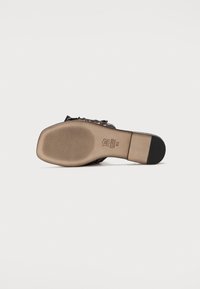 Sandale en cuir avec semelle plate, de couleur noire et marron, avec une texture antidérapante, design ouvert, détail de nœud sur le côté et bout arrondi.