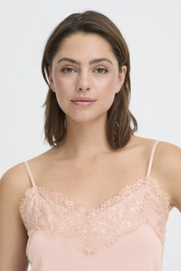 Mlada ženska s svetlo rjavimi lasmi, oblečena v bledo rožnato majico s čipkastim robom, obrnjena naprej z nevtralnim izrazom na obrazu.